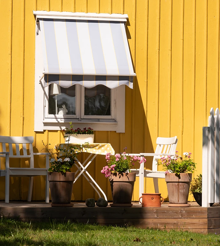 Gul träfasad med vita fönster, markis och blomkrukor på altanen