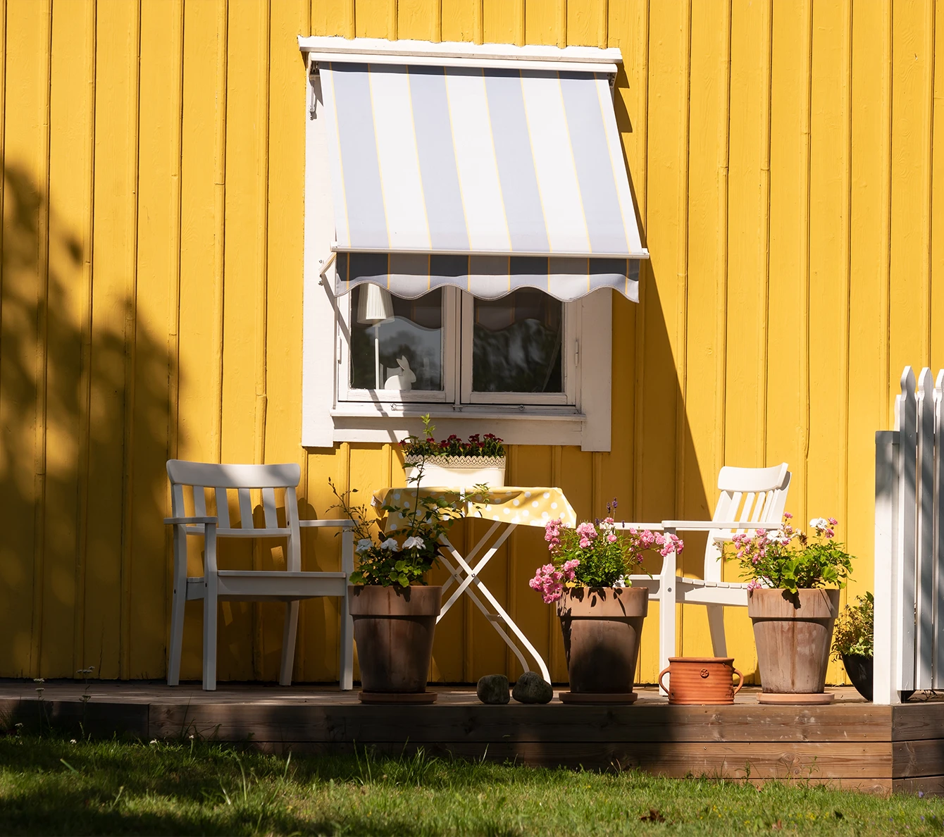 Gul träfasad med vita fönster, markis och blomkrukor på altanen